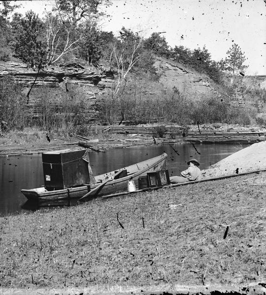 H.H. Bennett boat, with a dark tent and eighteen-by-twenty-two camera at a riverbank. An assistant is reclining on the edge of the river next to the boat.