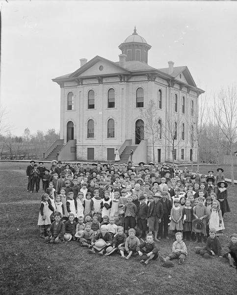 Slightly elevated view of an unidentified group of children and adults in front of a building. Possibly a school.