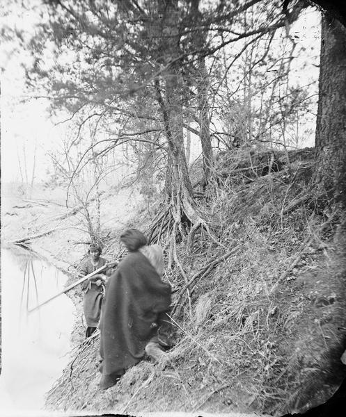 Two Ho-Chunk men are standing on a steep riverbank, with one aiming a gun.