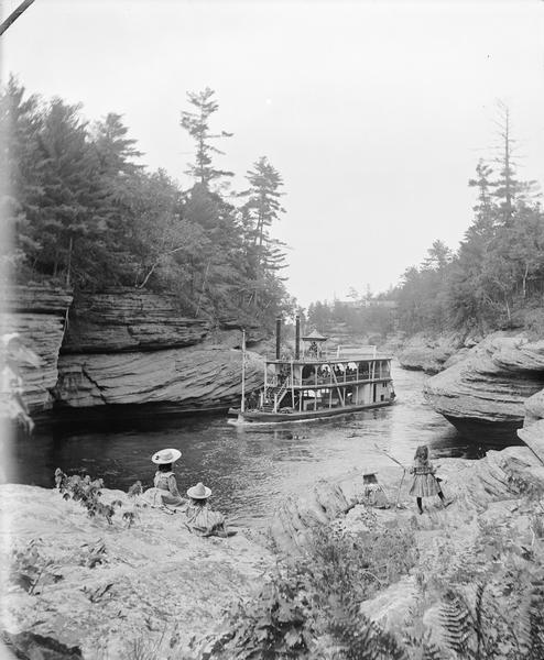 View down rock formation at Devil's Elbow towards the steamboat "Germania" at Narrows. Phyllis and Lois Crandall, and Miriam and Ruth Bennett girls are looking on from the rocky shoreline in the foreground.