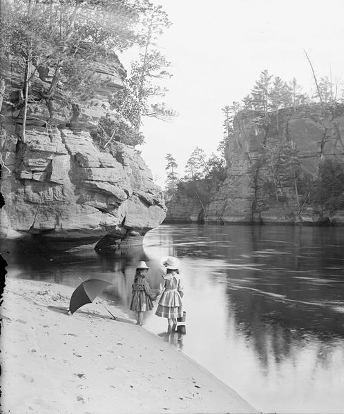 High Rock and Romance Cliff from bar above. Miriam and Ruth Bennett are standing at the sandy edge of the river holding hands. An open umbrella is laying next to them.