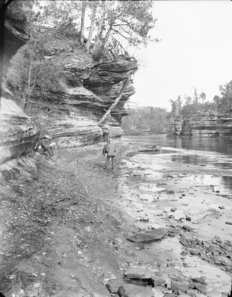 H.H. Bennett with camera near Steamboat Rock. There is a man on shore reading, and a woman in a canoe at the edge of the river.