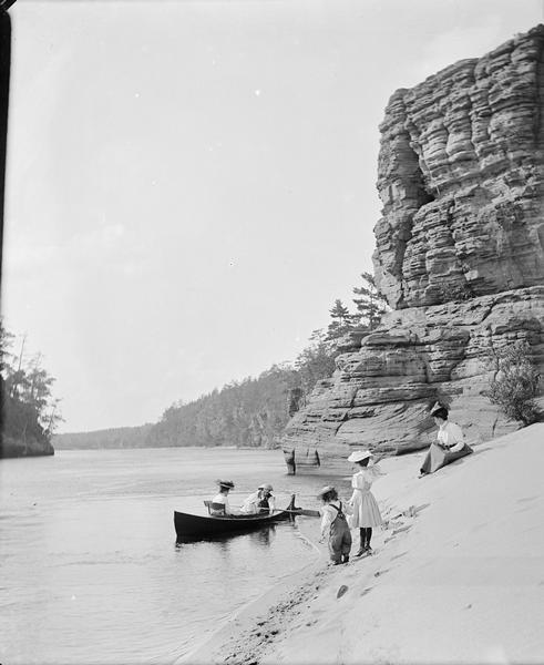 View along sandy shoreline towards the Bennett family on the beach at High Rock. Ashley and Helen are in the boat, and Miriam and Ruth are standing on the beach. Mrs. Bennett is sitting on the beach.