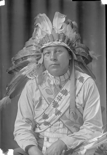 Waist-up studio portrait of Tom Walker wearing an eagle feather headdress and indigenous clothing.