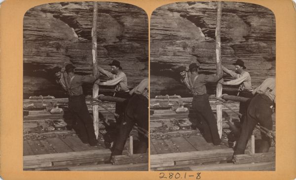 Stereograph of two men stabilizing a vertical log while two other men stabilize a horizontal log. Around both logs there is a rope rigged to hold the two logs together.