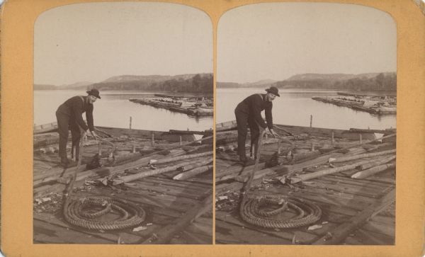 Stereograph of a man as he rigs a rope around a log on a lumber raft.