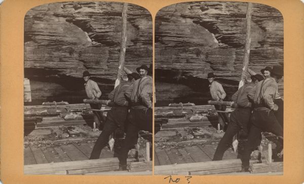 Stereograph of a group of men on a lumber raft holding a log vertical while other men push another log around it, creating a make-shift drill.