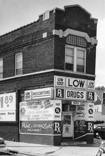 Exterior view of a pharmacy advertising "Low Cost Drugs." The building also has a painted advertisement for Pabst Blue Ribbon beer.