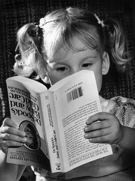 Young girl holding a book about child care written by Dr. Benjamin Spock, M.D. She is holding the book upside down.