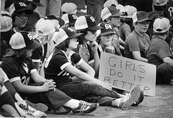 Young female baseball/softball team and young boys of opposing team sit on the sidelines. The girls are holding a sign that reads "Girls do it better."