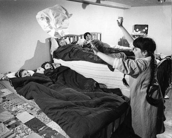 Young female children celebrate a sleepover while four girls in bed watch another girl toss a pillow in the air. They are smiling gleefully while the pillow floats in the air above them.