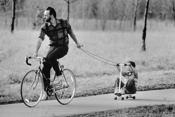 Father on a bicycle towing son on a scooter.