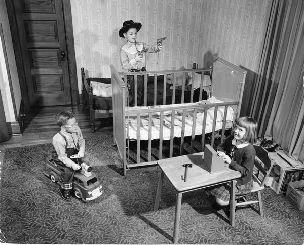 Elevated view of boy sitting on a toy fire engine, another boy dressed as a cowboy, and a girl with a toy cash register playing in the bedroom.