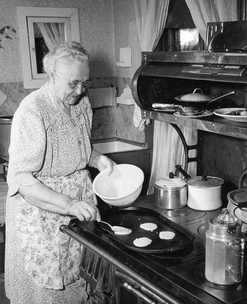 Elderly woman making pancakes on griddle at wood burning stove.