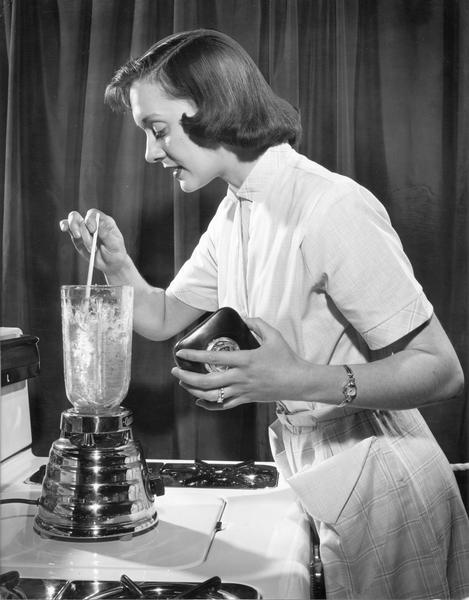 Woman stirring the contents of an Osterizer blender that is sitting on the stove.