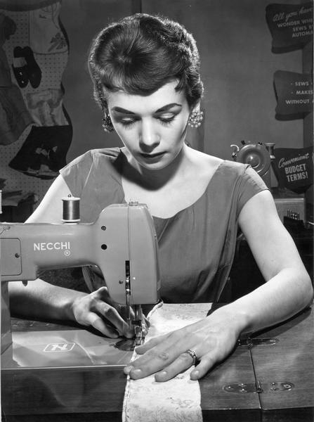 Woman using a Necchi sewing machine. Advertisements are in the background.