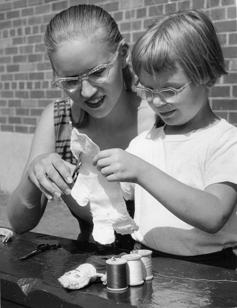 A young woman helps a girl make toy dolls out of cloth.