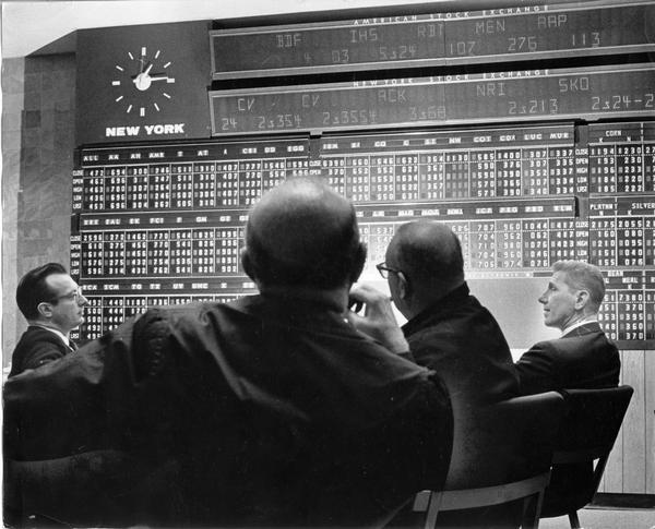A group of businessmen and customers watch a board that displays the stock amounts of the American Stock Exchange in a board room.