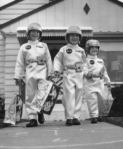 Three children, dressed up in homemade astronaut costumes for Halloween. One clever parent has made good use of a football helmet!