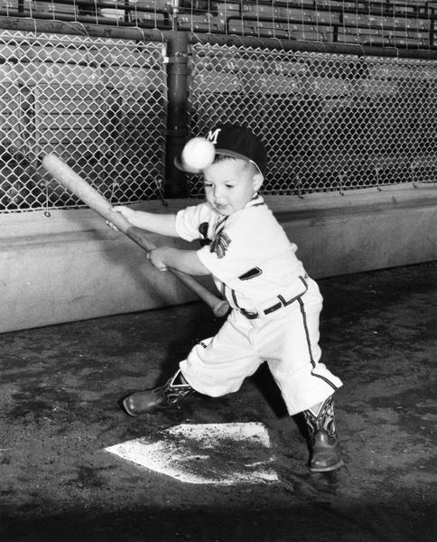 Three-year-old Edwin Lee Mathews, Jr., dressed in a "Milwaukee Braves" uniform, takes a swing at a baseball. He is the son of Braves' Hall of Fame third baseman Eddie Mathews.