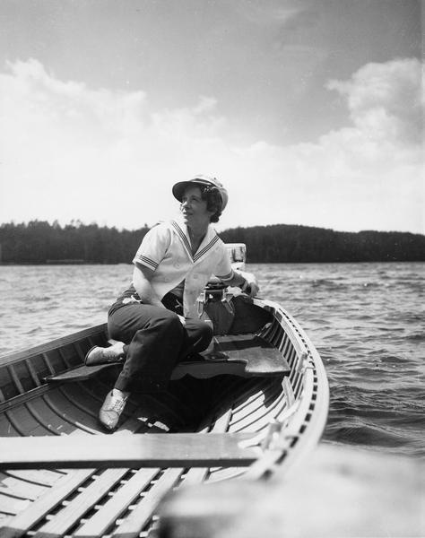 Woman at the tiller of a motorboat with tree-lined shore in the background. She is wearing a captain's hat at a jaunty angle and a sailor middy blouse.