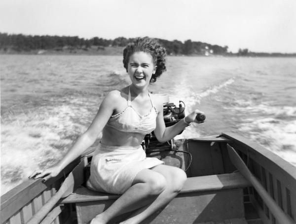 View of woman smiling and wearing a bathing suit while sitting in the back of a motorboat. She is steering the outboard motor on a lake. Behind her is the wake, and in the far background is a shoreline.