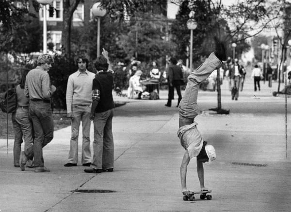 View down sidewalk of a skateboarder doing a handstand on a skateboard. Pedestrians are watching from the left.