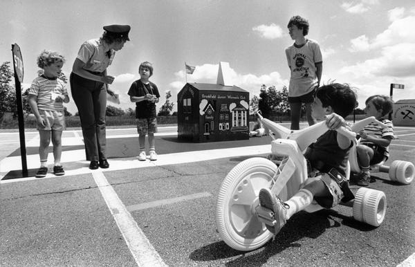 A group of children on "hot cycles," a form of young children's transportation, are being instructed on the rules of pedestrian and bicycle transportation safety.