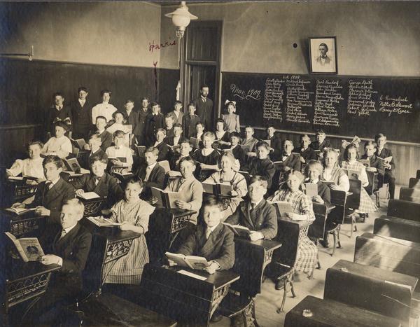 Slightly elevated view from front of students and teachers in an early 20th century school classroom. The names of all the students are written on the blackboard.