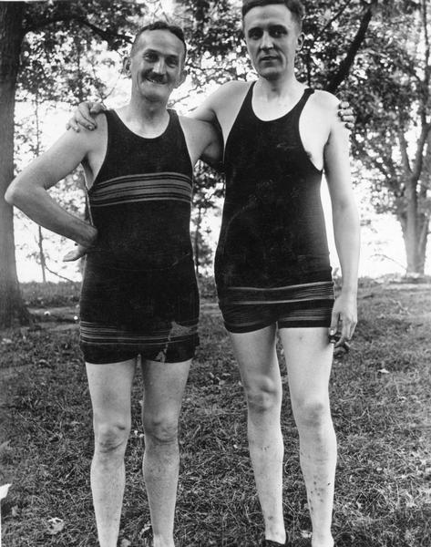Two men in bathing suits pose for portrait. They have their arms around each other and are smiling.