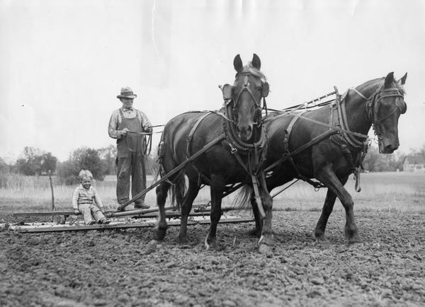 Farmer George Kopp giving a ride on his horse-drawn harrow to a young friend as he cultivates his field.