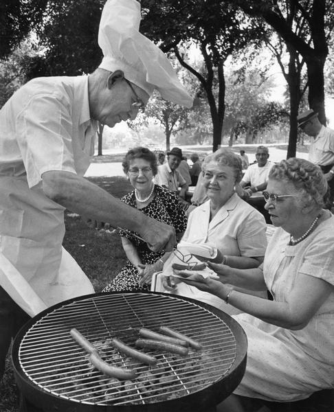 "Picnicking in Mitchell Park provided a pleasant afternoon recently for these senior citizens who participated in the Milwaukee County Park Commission's program for this age group. Chef for the day was Elmer C. Keller, 1960 S. 58th Street, who served barbecued frankfurters to other active members. They are Mrs. Kate Burmeister, left, 4103 S. 1st Pl.; Mrs. Theodore Witte, center, 2041 S. 33rd Street; and Mrs. Roy Langdon, right, 1424 W. Washington Street. All except Mrs. Langdon have contributed recipes to the 'Senior Citizens Cookbook'."