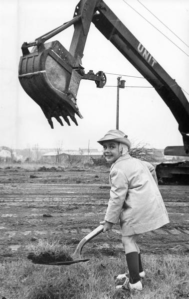 A three-year-old poses with a man-sized shovel of soil during a groundbreaking ceremony at the site of a new preschool. Behind him is a large steam shovel.