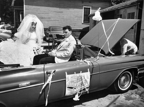 Bride and groom wait in the open convertible that is their transportation to their reception while a mechanic works under the hood.