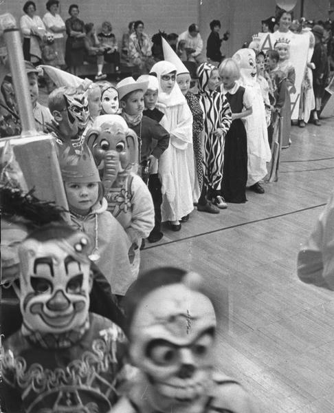 A line of elementary students dressed up in Halloween costumes at school. Women are standing and sitting in the background.