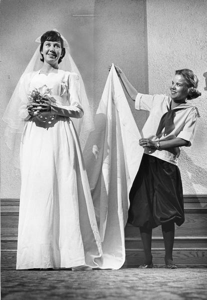 Two women model clothing. The woman on the left is wearing a wedding dress from 1946, and the other woman is wearing a Mount Mary gym suit.