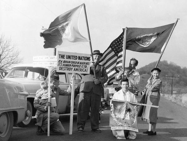 Glenn Turner is holding the United Nations flag, and his wife is carrying the flag of the state of Wisconsin. A supporter is holding the American flag. One man is kneeling on the road and holding a young child on his arm while holding onto two signs. One reads: "Get the U.N. Out of [obscured] Get the U.S. Out of [obscured]", and the other reads: "THE UNITED NATIONS: Is a brain child of Alger Hiss it's hidden purpose it to ... DESTROY AMERICA!" A woman kneeling in front of the group is holding a banner depicting a spider in a web with the words: "Won't you come into my parlor?" The group had encountered a roadblock and protesters are opposing the United Nations. A person obscured by the American flag is holding a camera and standing just behind the group.