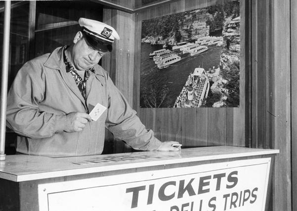 A man operating a ticket booth in the Wisconsin Dells. He is playing a card game, probably solitaire, on the counter.
