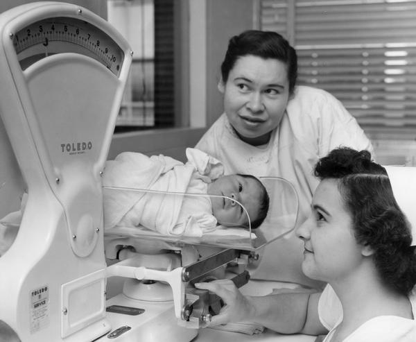 A new mother is looking on as her 13-pound newborn infant is weighed by the nurse.
