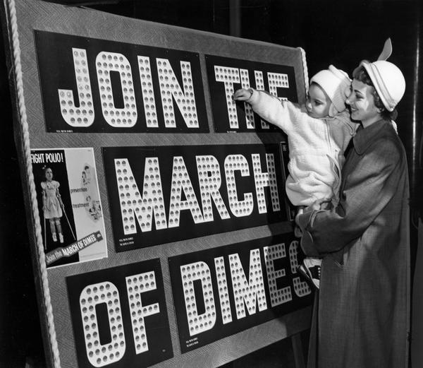 A toddler is inserting a dime into a March of Dimes collection board as her mother is holding her in her arms and looking on. The collection is to raise money to eradicate polio.