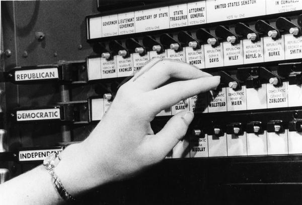 Close-up view of a women's hand touching a ballot machine with names of candidates from the 1956 Wisconsin election.