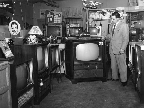 A man standing in a television retail store. A number of television sets are displayed, including one Hallicrafters set.