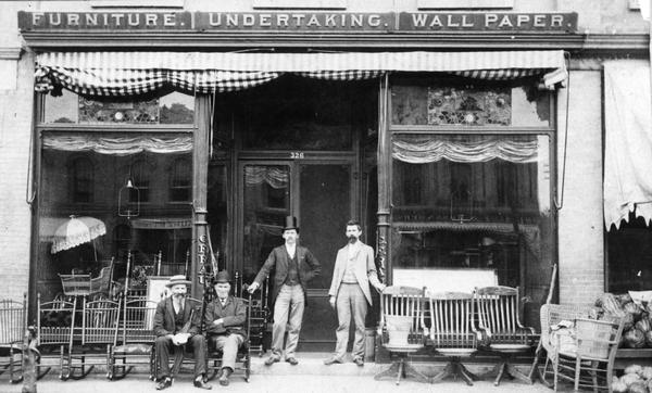 Front view of C.F. Rau's furniture store, with the owners and employees sitting in front of the store. Apparently, the store also performs undertaking. The location is unknown but it is assumed to be either Rockford, Ill., or Beloit, WI. C.F. Rau and "Mr. Dow" are standing in the doorway; two (visitors, unnamed) are seated in chairs on display along the street; next door is Charles Greene's grocery.
