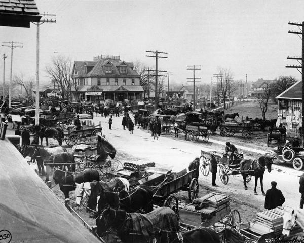 Elevated view of the monthly Stock Fair. The roof of a building is in the left foreground. Horse-drawn wagons line the street, and there is a large building at an intersection further down the street where a large group of people are gathered.
