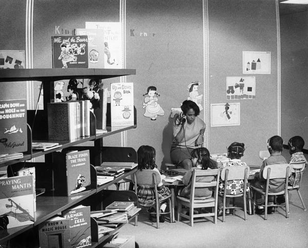 Young children listen to a story through earphones as a teacher's aid looks on.