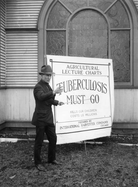 Man appearing to deliver a lecture on tuberculosis prepared by International Harvester's Agricultural Extension Department. The man is pointing to an "agricultural lecture chart" and is standing in front of Big Woods Congregational Church.