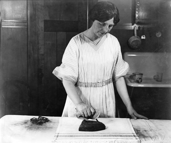 Woman ironing cloth with old-fashioned heated iron. Behind her is a sink.