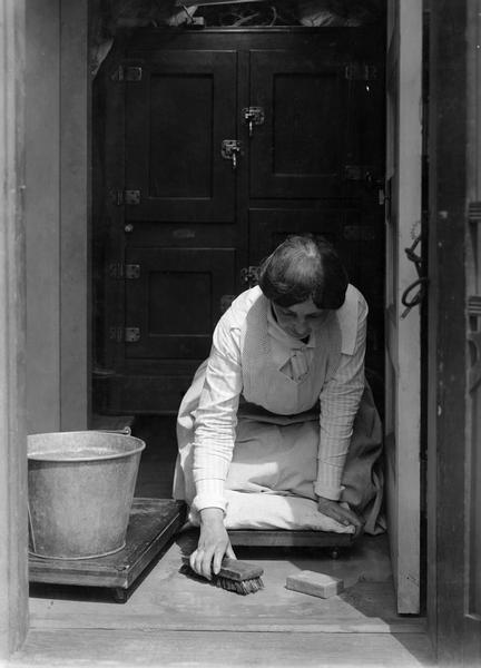 Woman cleaning a wooden floor in an open doorway of her farmhouse with a scrub brush.