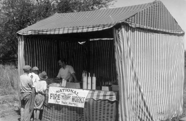Three boys purchase fireworks from the proprietor of a National Fireworks tent near Ogden Avenue. The photograph was taken for International Harvester's Agricultural Extension Department to caution families against potential farm hazards.