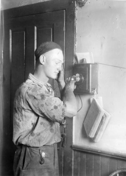 Man using a telephone mounted to an interior wall. The photograph was taken for International Harvester's Agricultural Extension Department. It was used to warn families of potential farm hazards. The original caption reads: "Don't talk on a telephone during an electric storm."